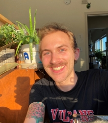 Adam Mastel - short blonde hair, mustache, black t-shirt, houseplant in background